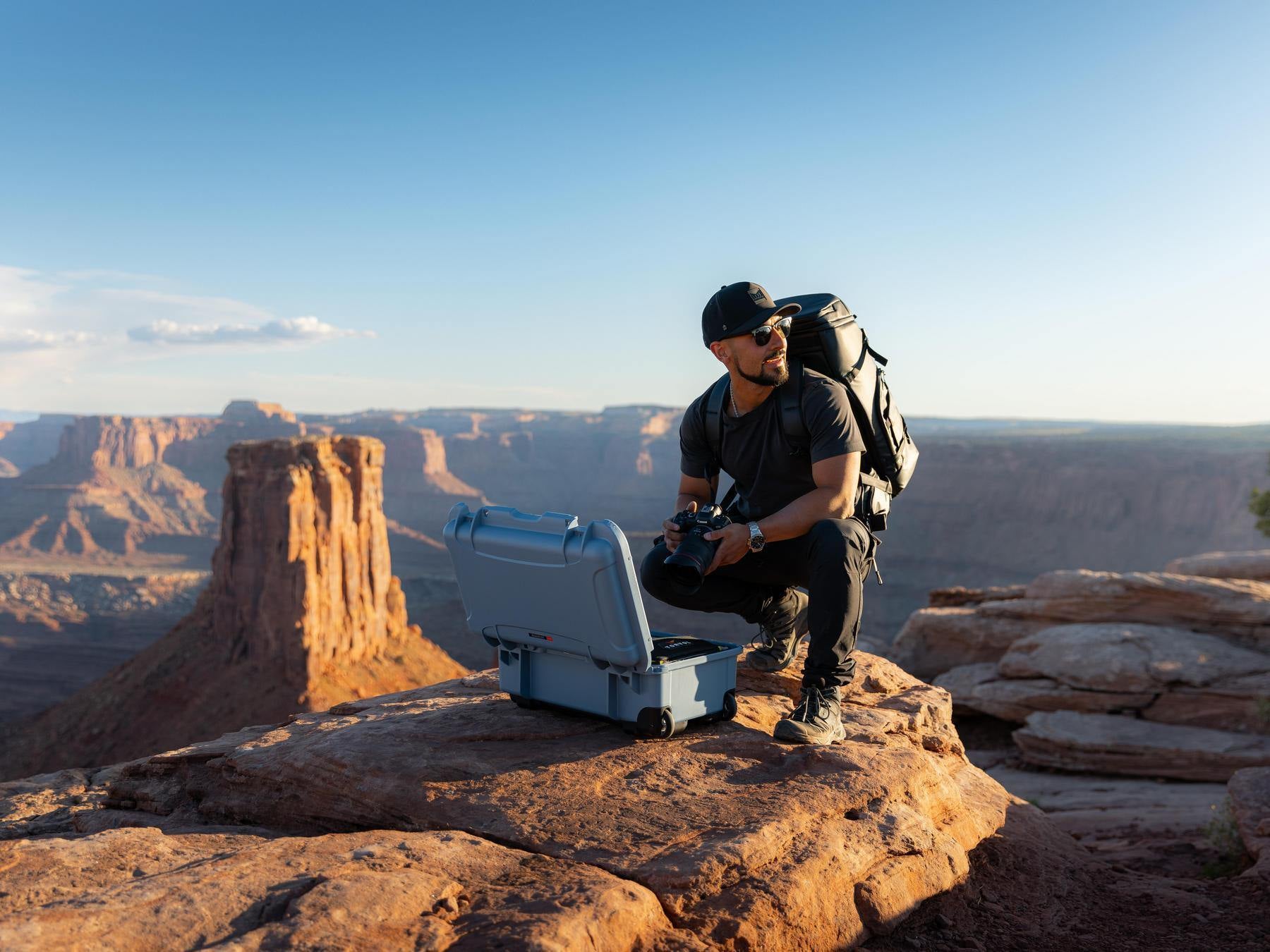 Person with a backpack and camera equipment sitting on a rocky outcrop with a scenic landscape in the background.