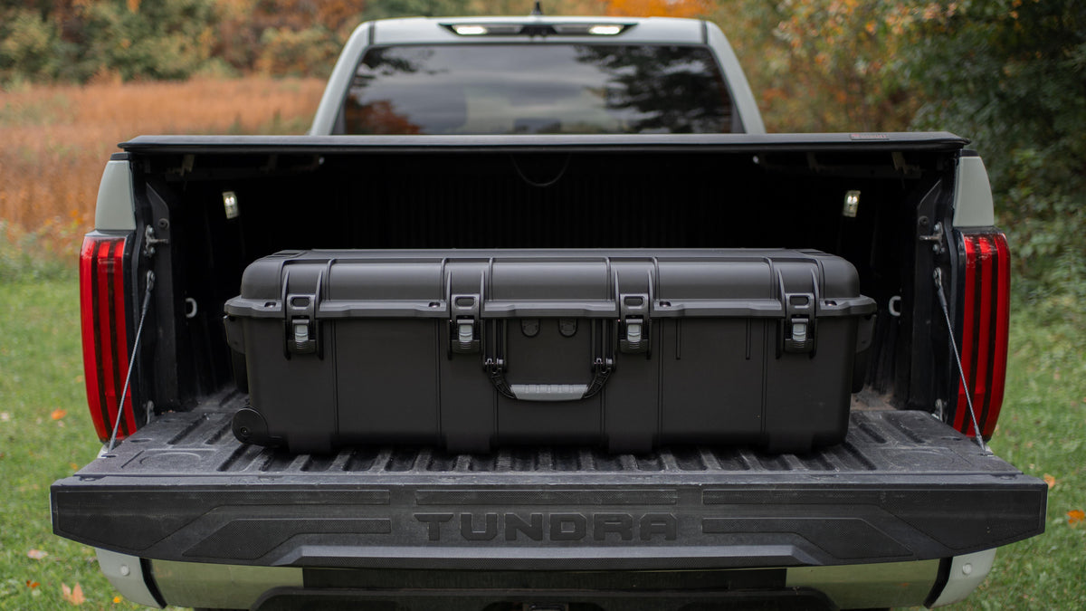 Un grand valise e de rangement noir à coque rigide chargé sur le plateau d'un pick-up argenté, vu de l'arrière avec le hayon abaissé. Le pick-up est garé à l'extérieur sur de l'herbe, avec des arbres en arrière-plan.