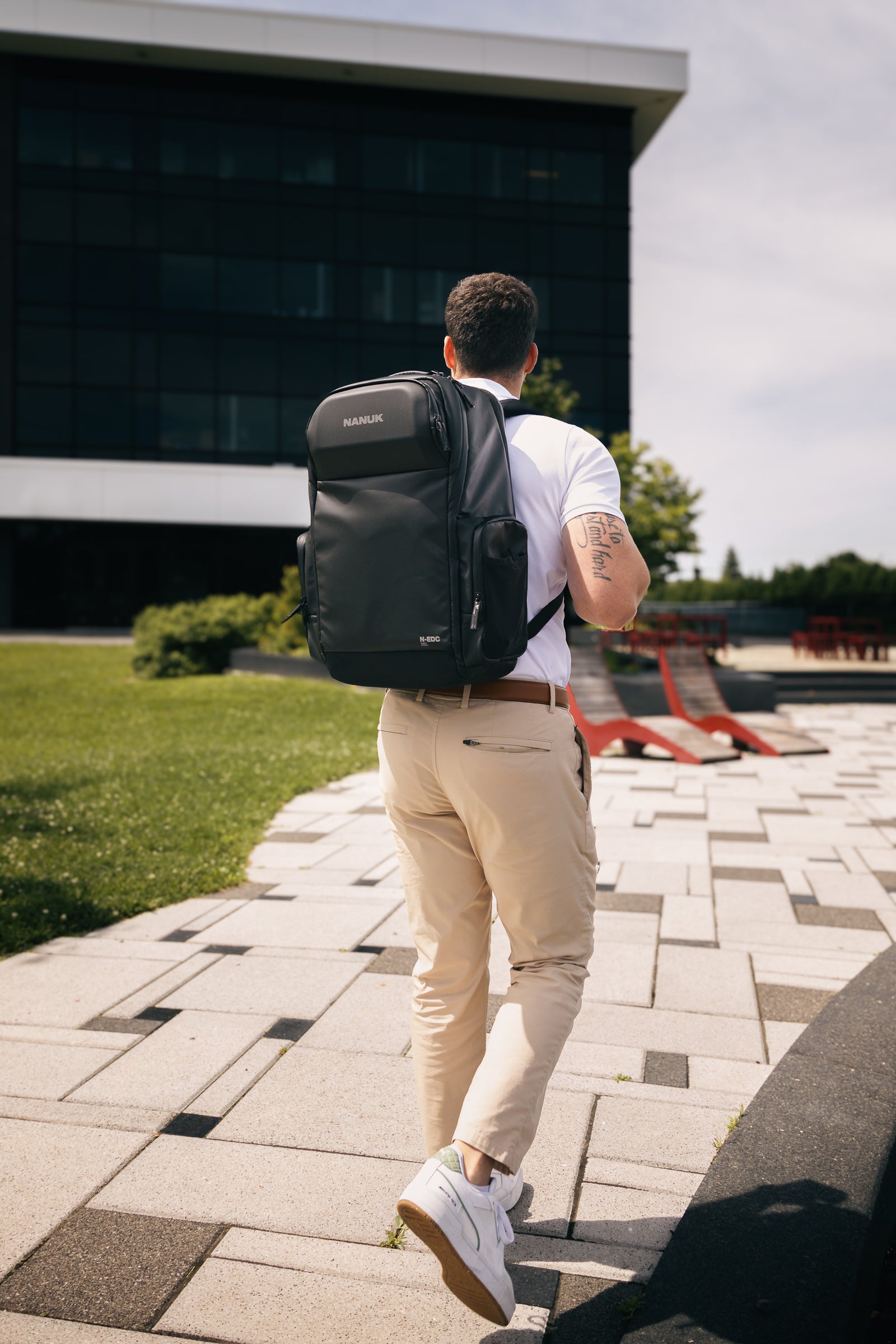 Mann, der mit einem schwarzen Rucksack im Freien spazieren geht, bekleidet mit einem weißen Hemd und einer beigen Hose.