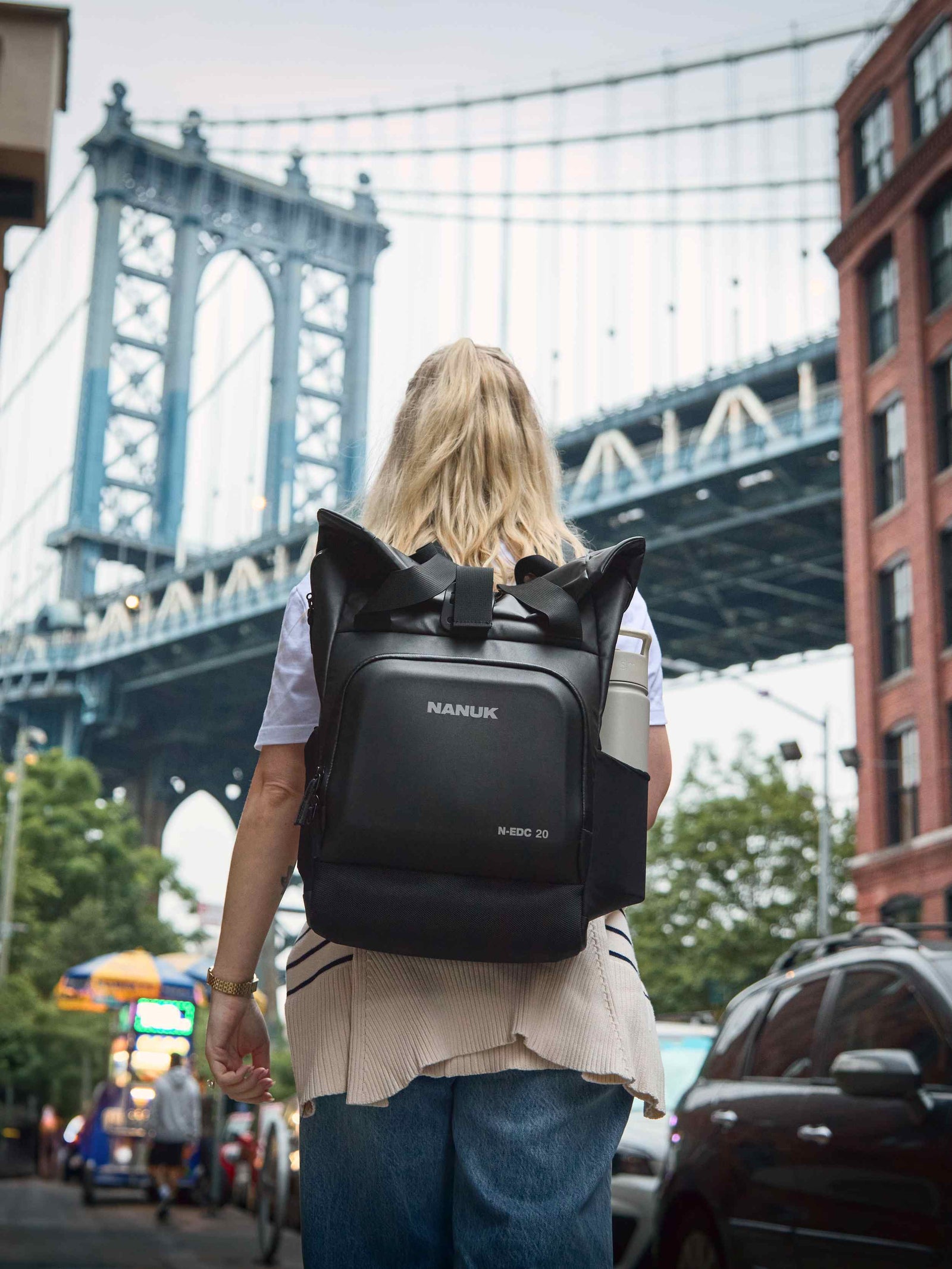 Person mit einem Nanuk-Rucksack, die in einer städtischen Umgebung mit einer Brücke im Hintergrund spazieren geht.