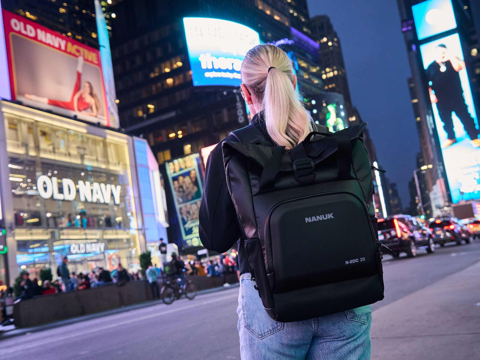 Person mit einem schwarzen Rucksack, die nachts durch eine belebte Stadtstraße läuft.