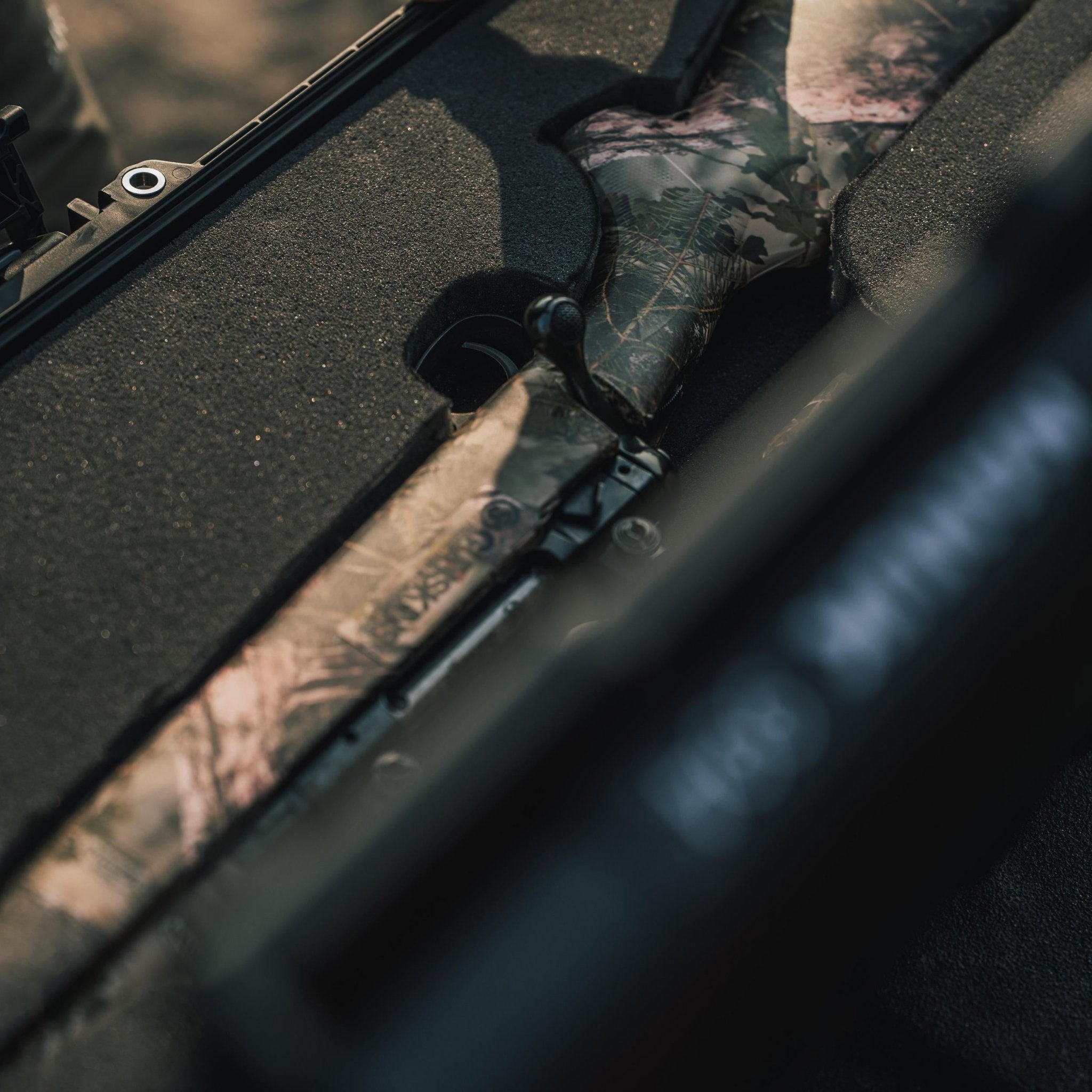 Gewehr mit Tarnmuster in einem Fahrzeug, mit Fokus auf dem Gewehrgriff.
