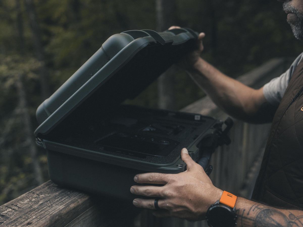 A man outdoors on a wooden deck by a forest, opening a dark green hard protective case. The man is wearing a brown vest and a smartwatch with an orange band.