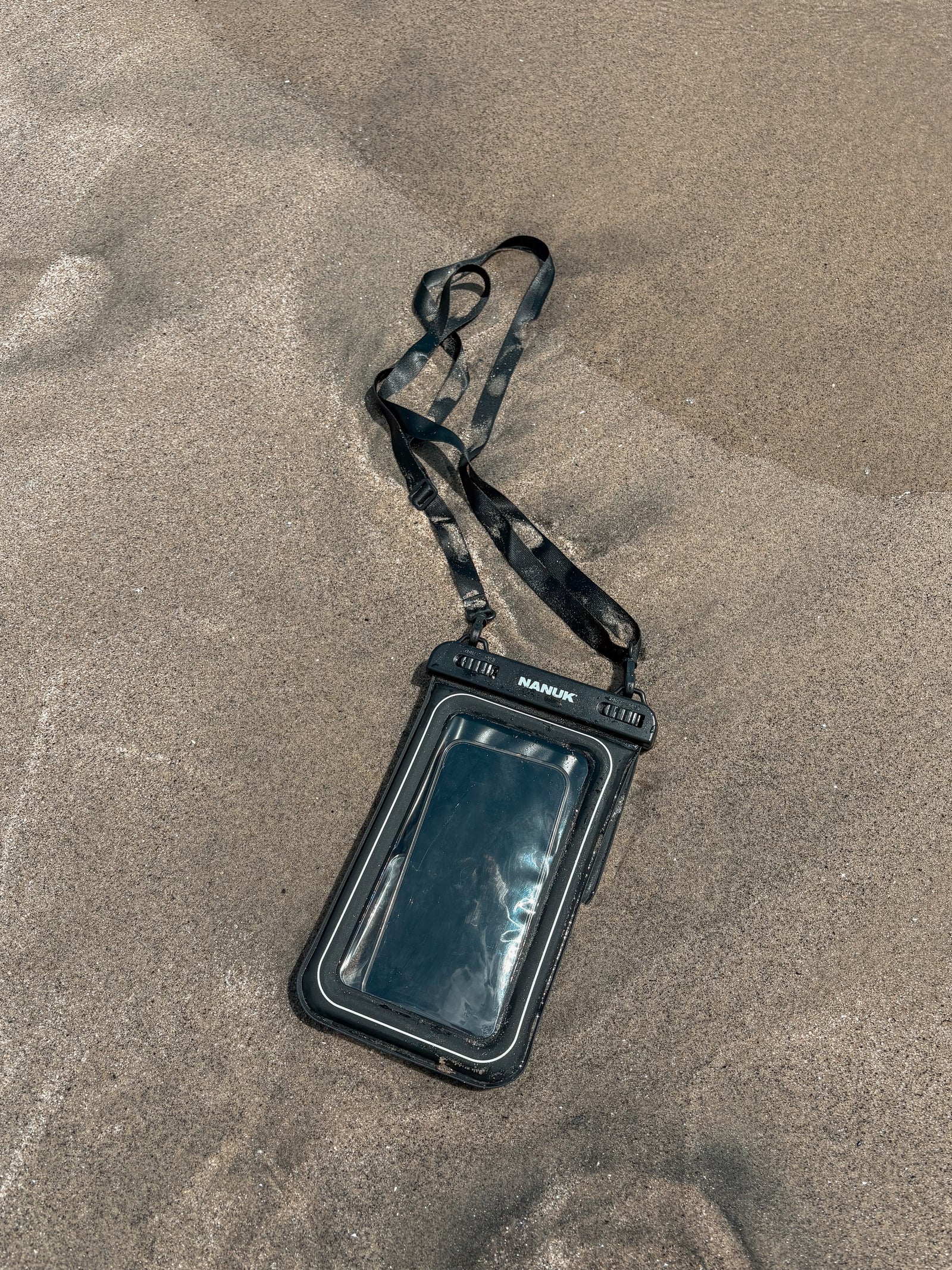 Eine schwarze, wasserdichte Handytasche mit einem durchsichtigen Sichtfenster und einem dazu passenden, schwarz gemusterten Trageband, die auf sandigem Boden liegt.
