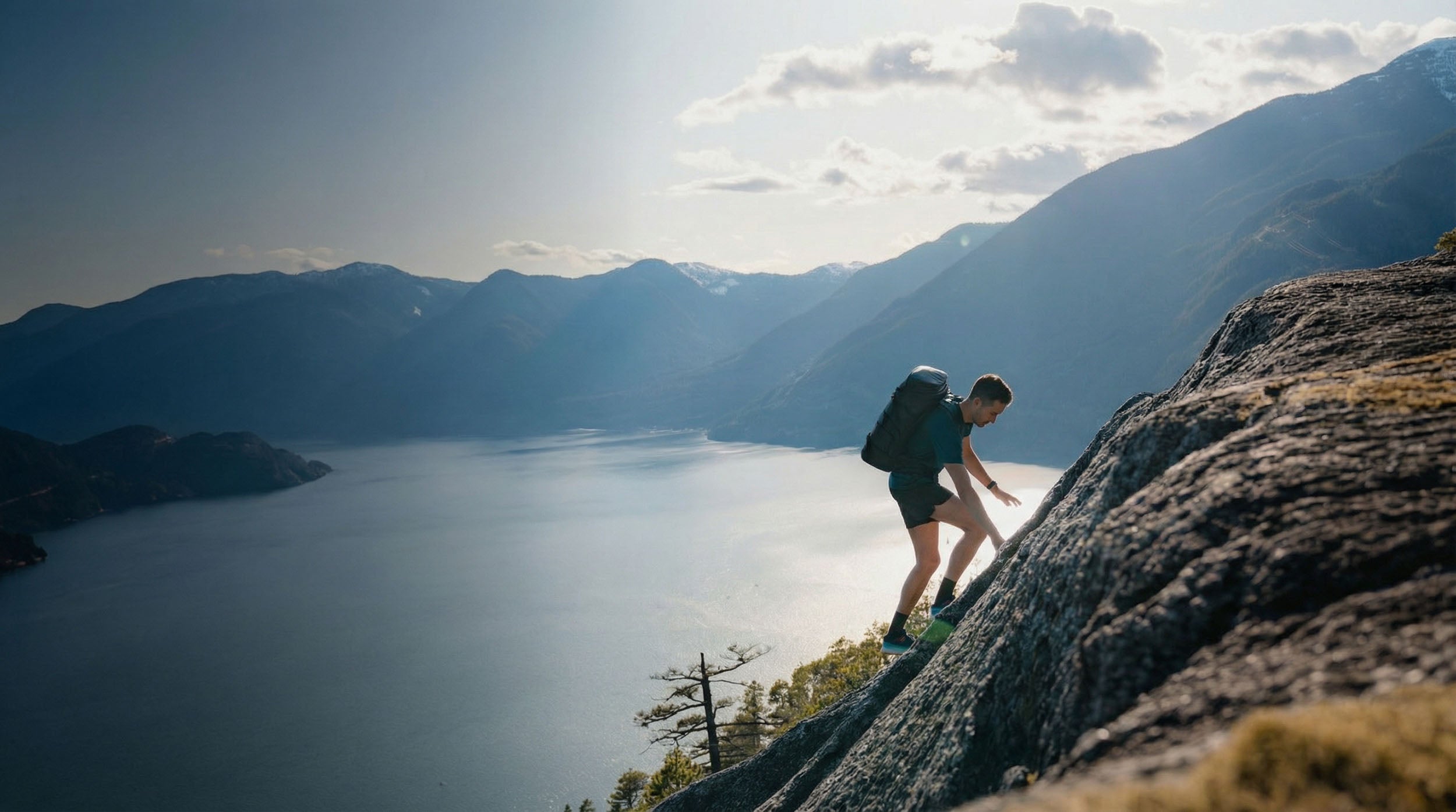 A hiker with a backpack scrambles up a steep rocky mountain ridge overlooking a scenic lake and misty blue mountains.