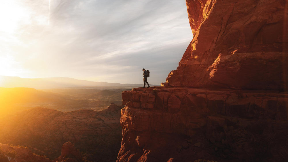 Person standing on a rocky outcrop overlooking a vast landscape with a sunset.