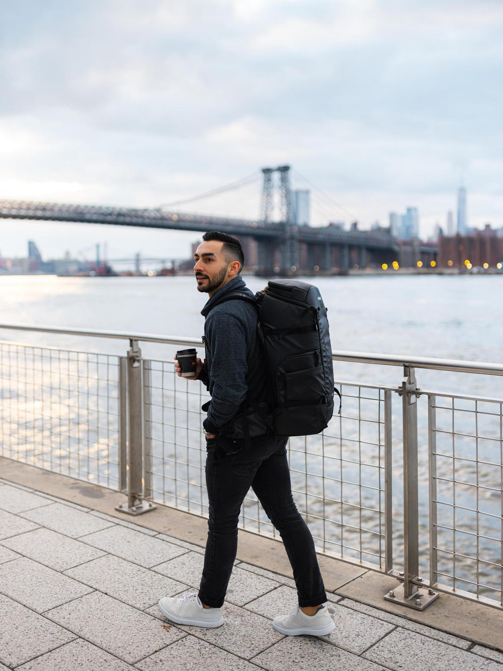 Man with a backpack walking by a waterfront with a bridge in the background