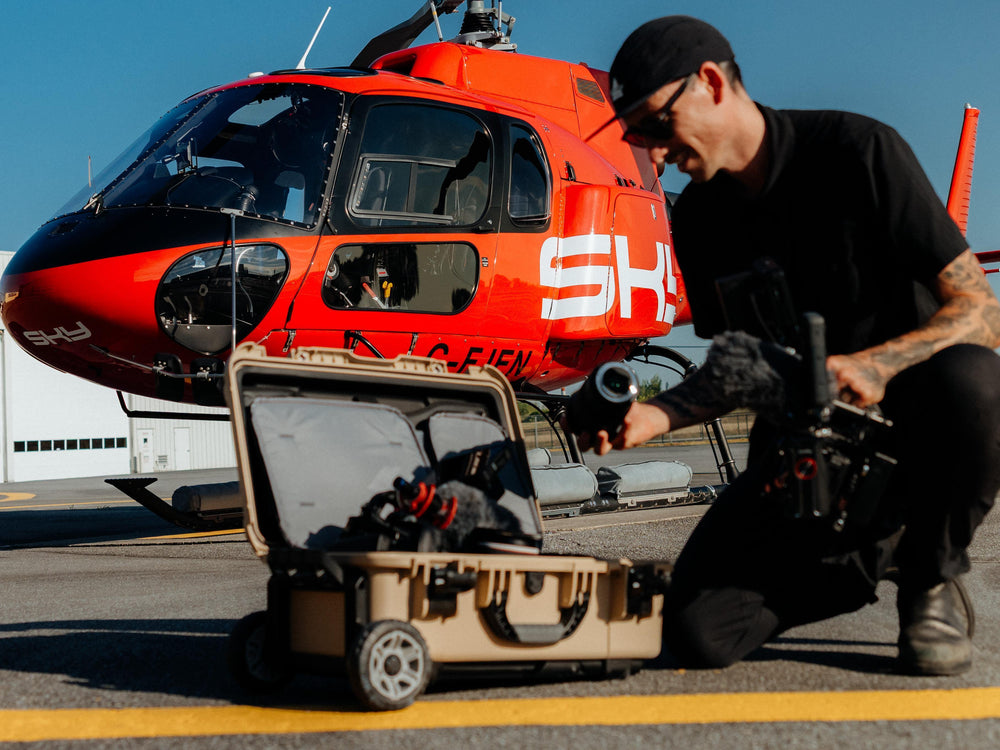 Ein Mann in schwarzer Kleidung kniet auf dem Rollfeld eines Flughafens vor einem leuchtend roten Hubschrauber und hält einen Kamerasteuergerät neben einem offenen, beigen Hartschalenkoffer, in dem sich eine Drohne befindet.