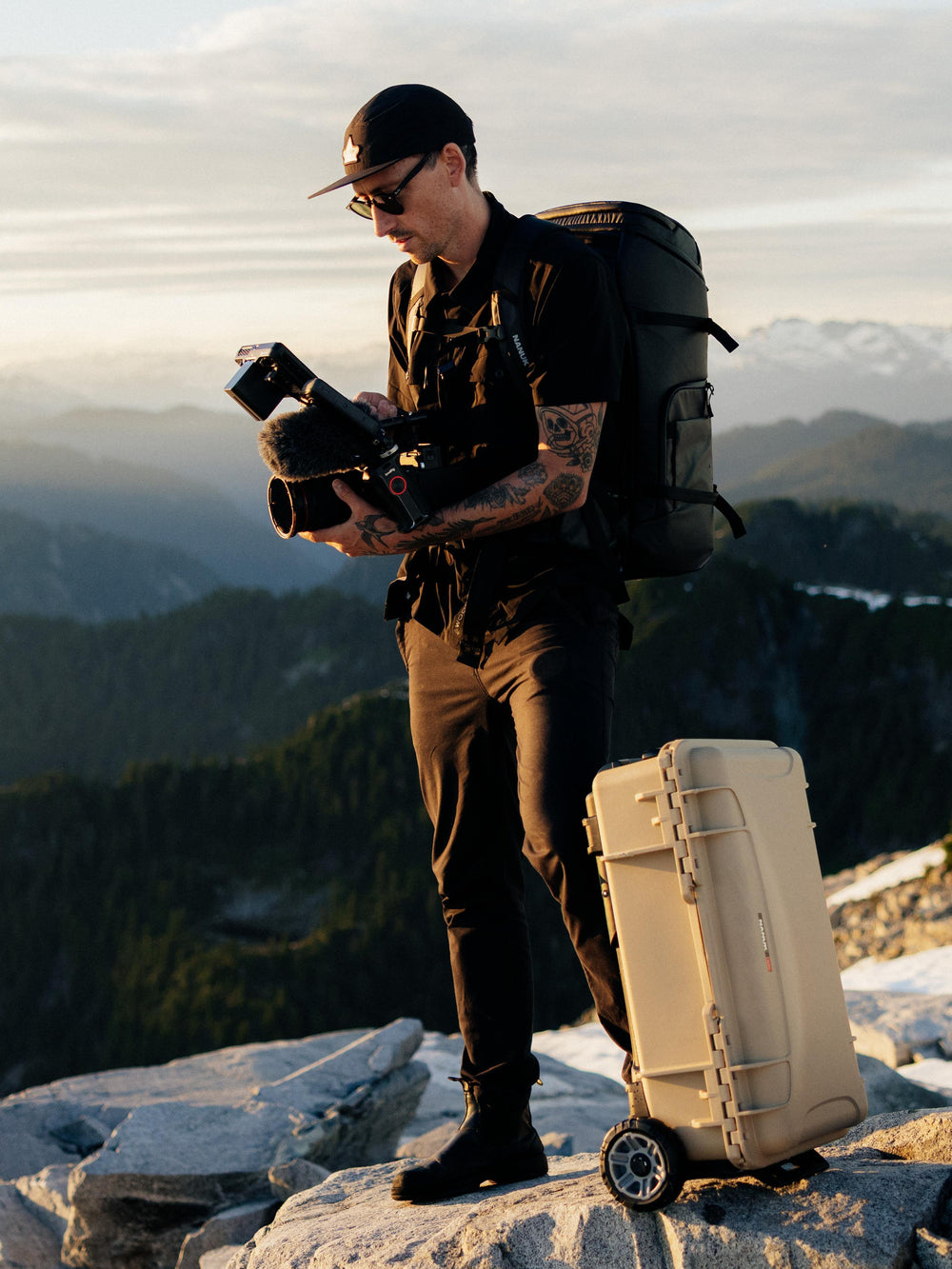 Ein Mann steht mit einer Kamera und einem großen schwarzen Rucksack auf einem Berggipfel, neben ihm steht ein beiger Hartschalen-Rollkoffer. Im Hintergrund ist eine malerische Berglandschaft zur goldenen Stunde zu sehen.