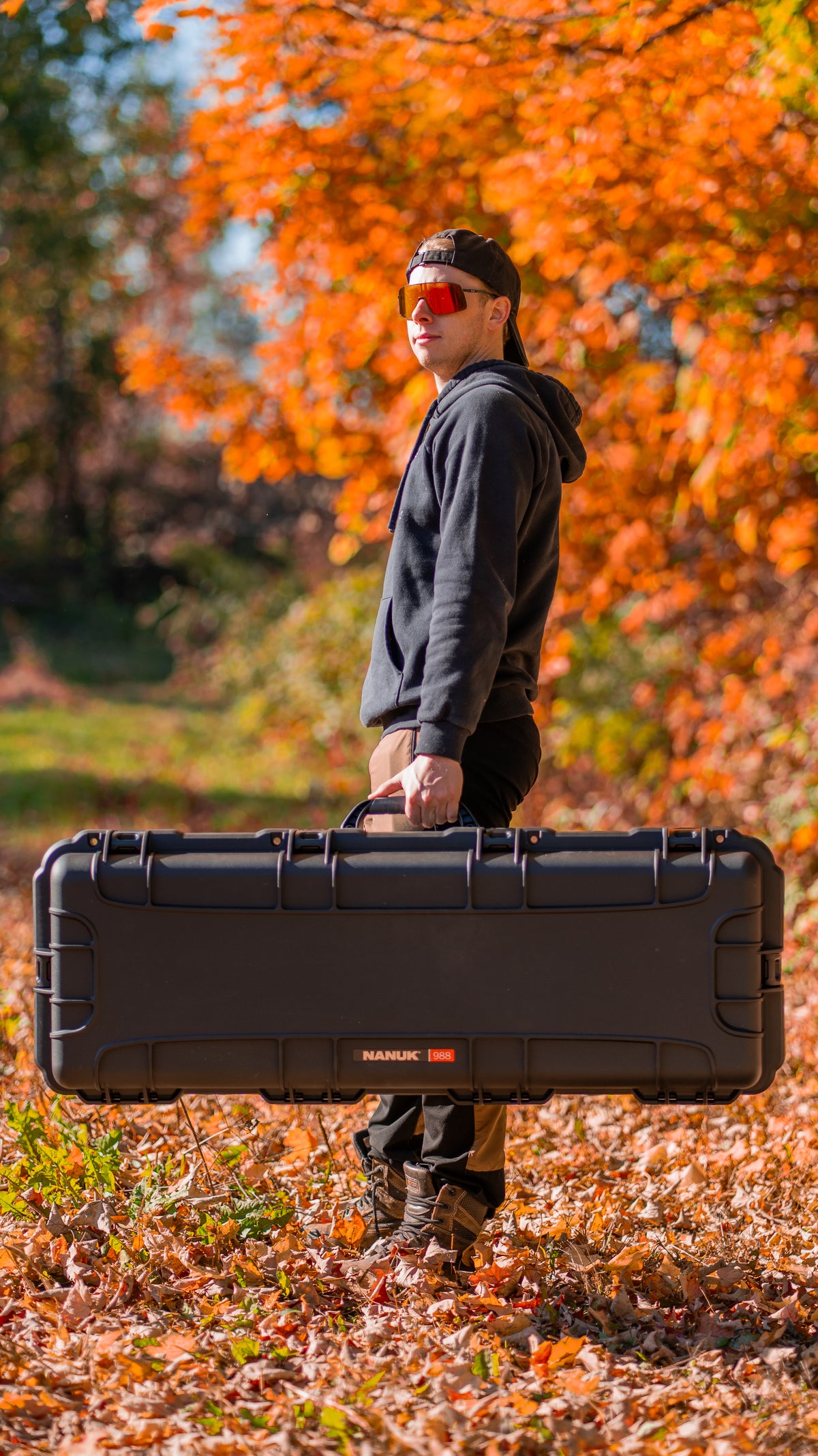 Un homme vêtu d'un sweat à capuche noir, d'une casquette noire et de lunettes de soleil aux verres teintés d'orange transporte une grande valise rigide noire de type « valise » à l'extérieur, dans un décor automnal où l'on aperçoit des feuilles orange au sol et des arbres en arrière-plan. La marque « MANUKE » est visible sur le devant de la valise « valise ».