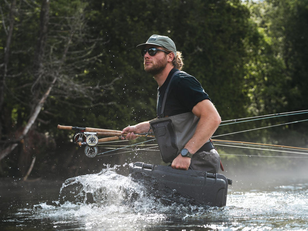 Man fishing in a river with waders and a fishing rod, surrounded by trees.