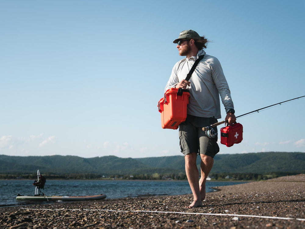 Man walking on a beach with a red cooler and fishing rod, wearing sunglasses and a cap.