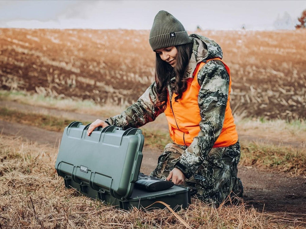 Person in camouflage jacket and orange vest opening a green hard-shell case in an outdoor setting.