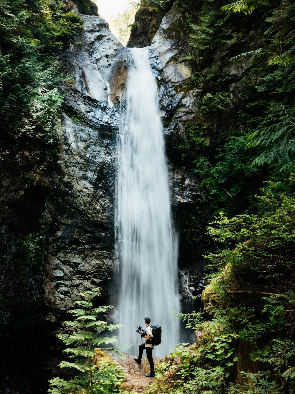 Ein hoher Wasserfall stürzt eine felsige Klippe hinab, die von dichtem, grünem Wald umgeben ist; am Fuße des Wasserfalls steht eine kleine Person mit einer Kamera in der Hand.