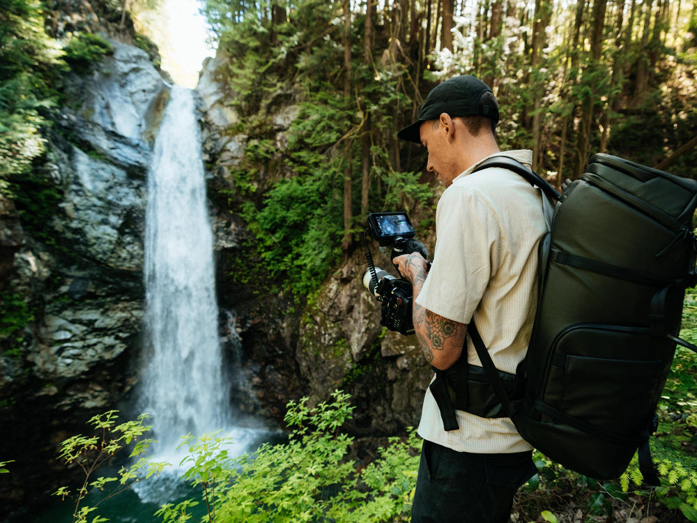 Ein Mann, der ein beiges T-Shirt, eine schwarze Mütze und einen großen schwarzen Rucksack trägt, steht in einem üppigen Wald neben einem Wasserfall und hält eine Kamera mit einem Bildschirm in der Hand.