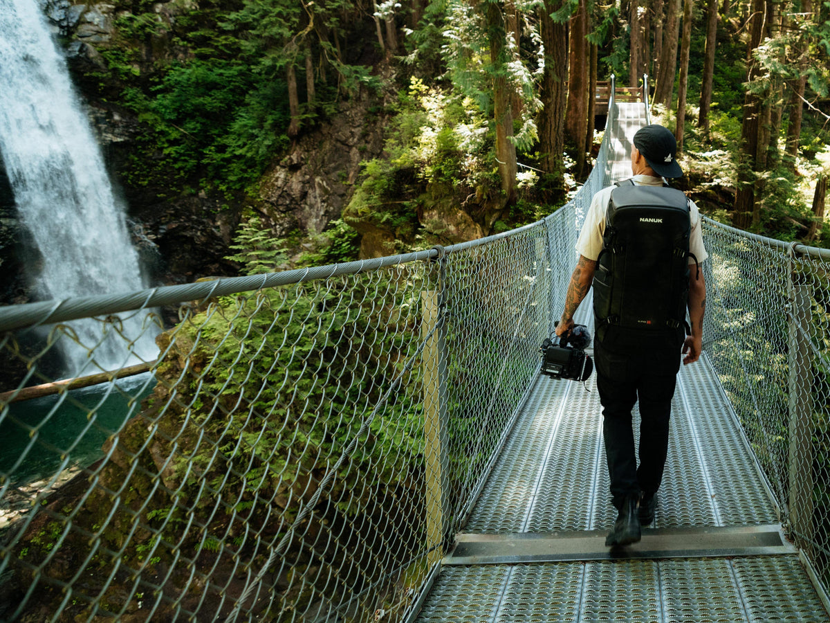 Ein Mann mit einem schwarzen Rucksack, der über eine Hängebrücke in einem Wald geht; auf der linken Seite ist ein Wasserfall zu sehen. Er trägt eine Kamera bei sich.
