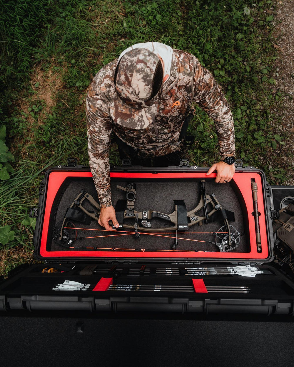 Person in camouflage gear setting up a bow in a black and red case outdoors.