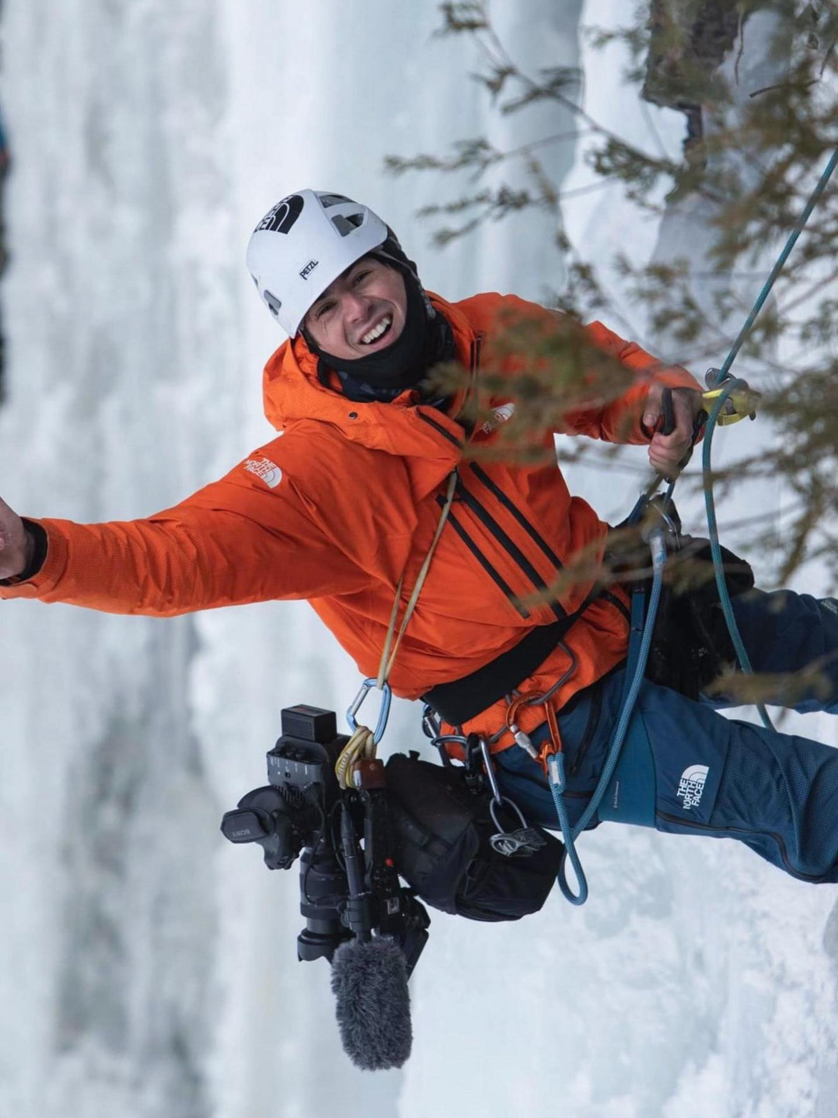 Person ice climbing in a snowy landscape wearing an orange jacket and helmet.