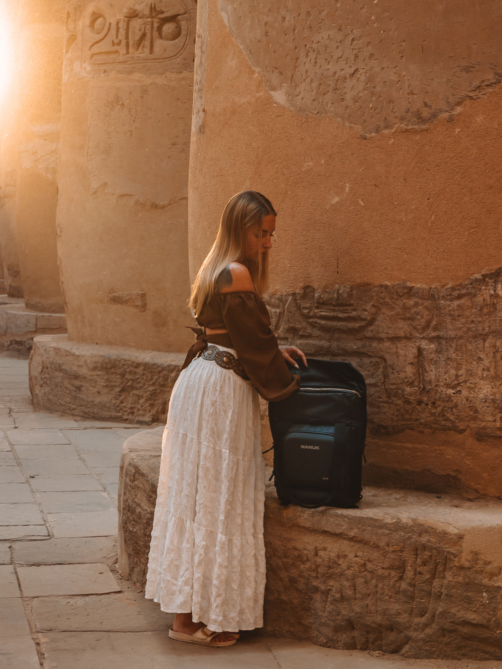 Woman in a long white skirt and brown top using a black suitcase in front of ancient stone walls.