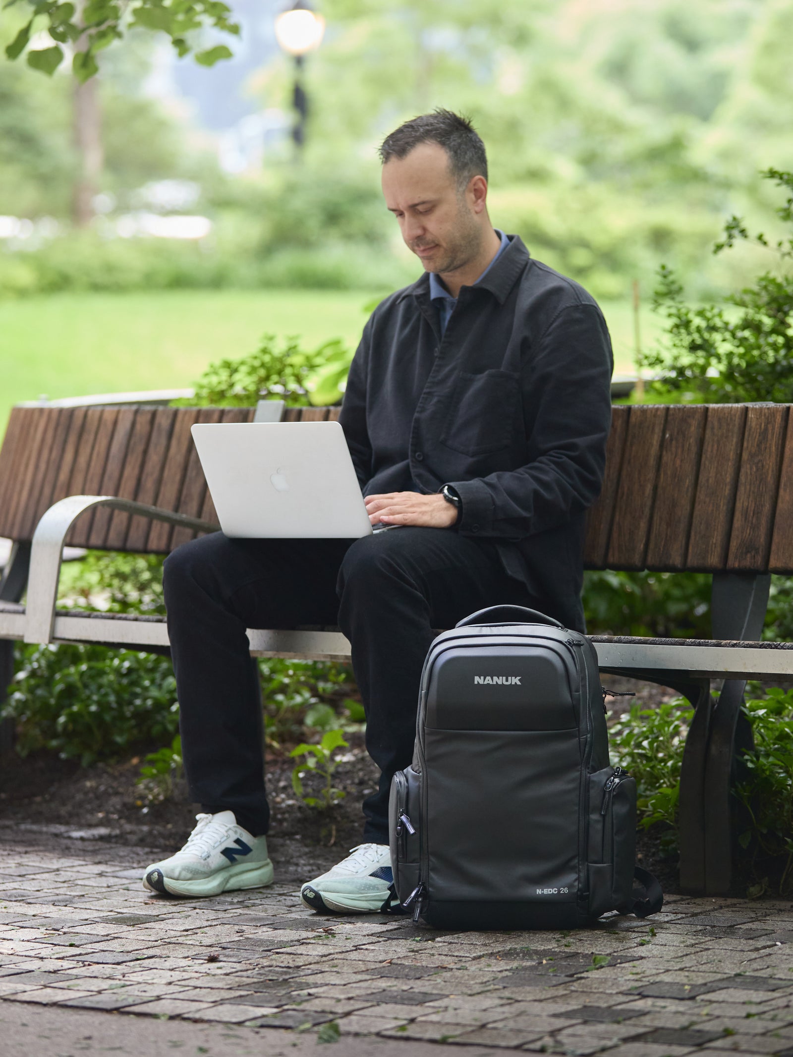 Mann sitzt auf einer Parkbank und benutzt einen Laptop, neben ihm steht ein Nanuk-Rucksack.