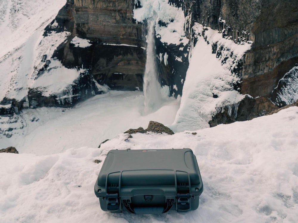 Black hard-shell suitcase on a snowy landscape with a waterfall and ice formations.