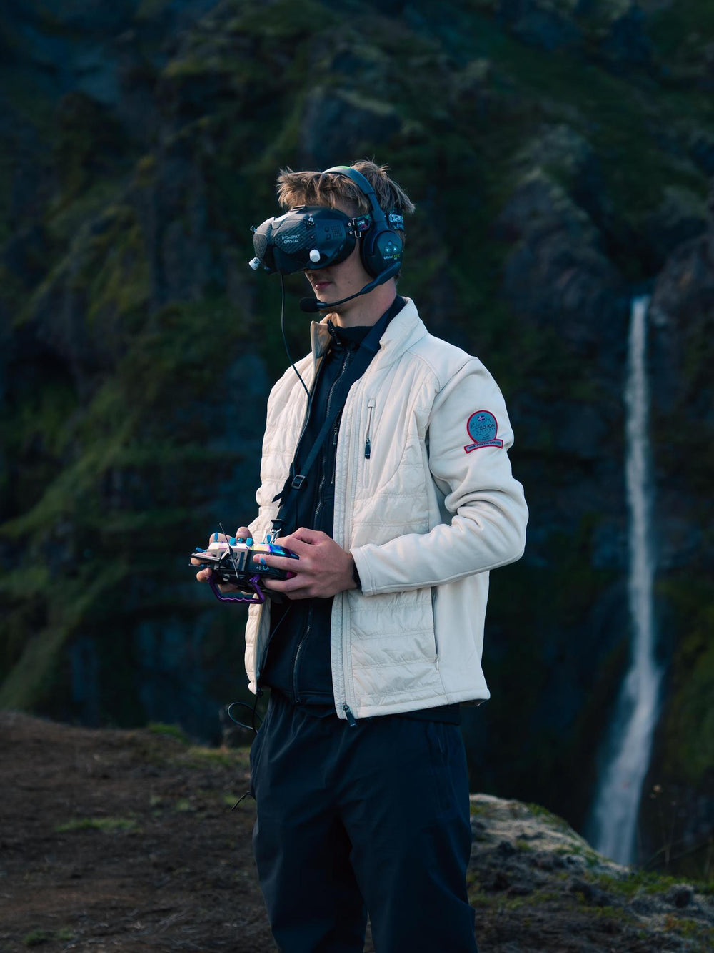 Person wearing VR goggles and holding controllers in a natural setting with a waterfall.