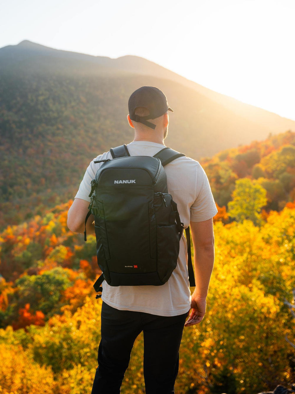 Person wearing a black backpack with 'NAMUK' branding, standing in a scenic outdoor setting with mountains and trees.