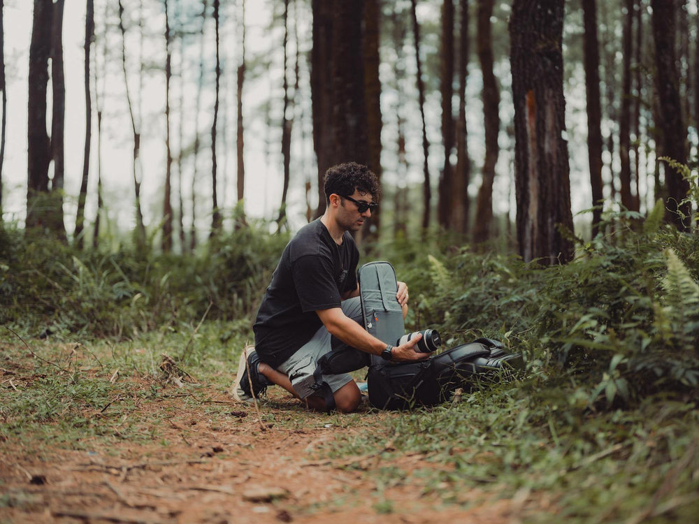 Man sitting in a forest with a backpack and camera, surrounded by trees and greenery.