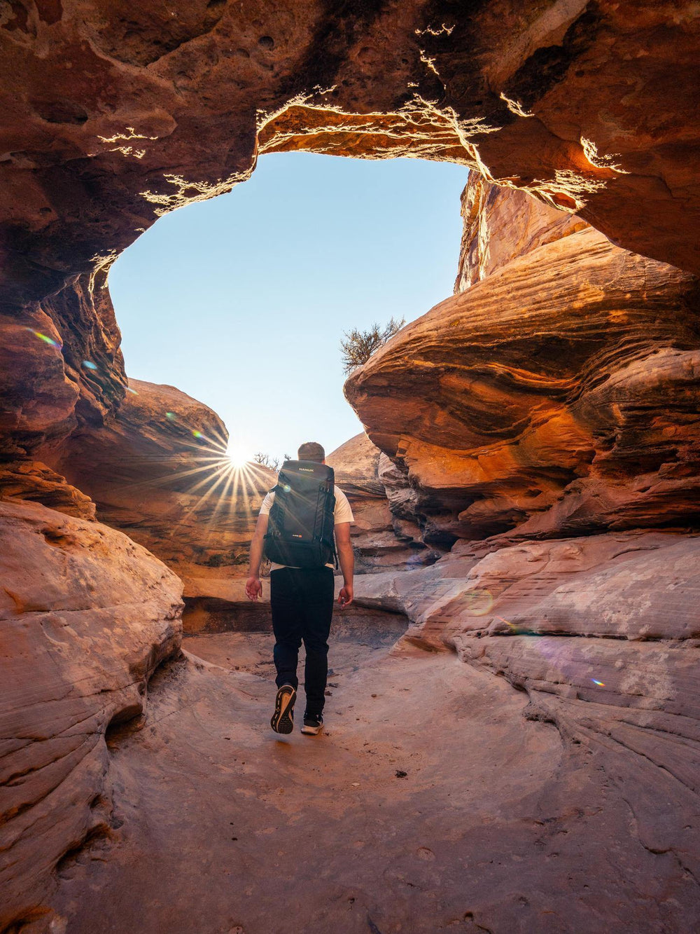 Person walking through a rocky archway with a backpack, sun shining through