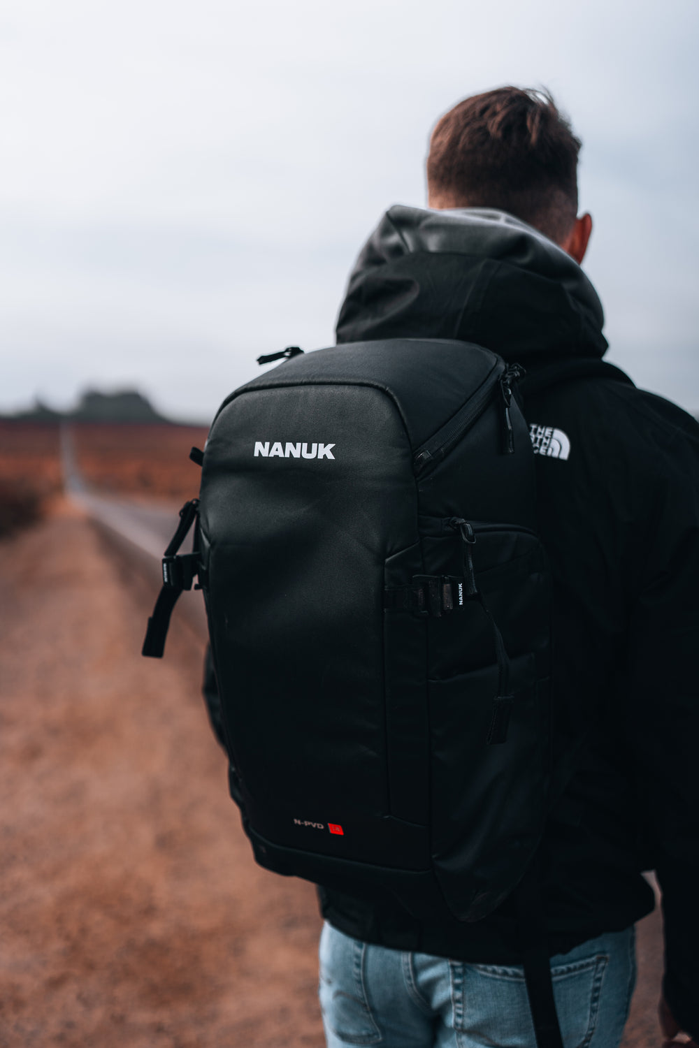 Person wearing a black Nanuk backpack with The North Face logo, standing on a path.