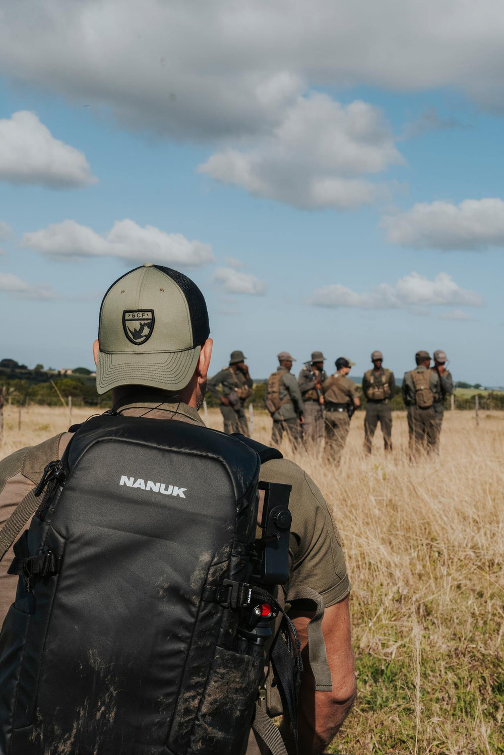 Person with a Nanuk backpack observing a group of people in military-style gear in a field.