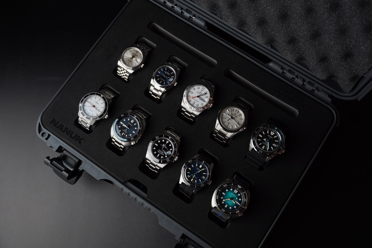 An open black hard case with a foam insert holding ten wristwatches, photographed on a dark studio background. The watches have mixed dial colors including white, blue, and green, with silver and black cases.