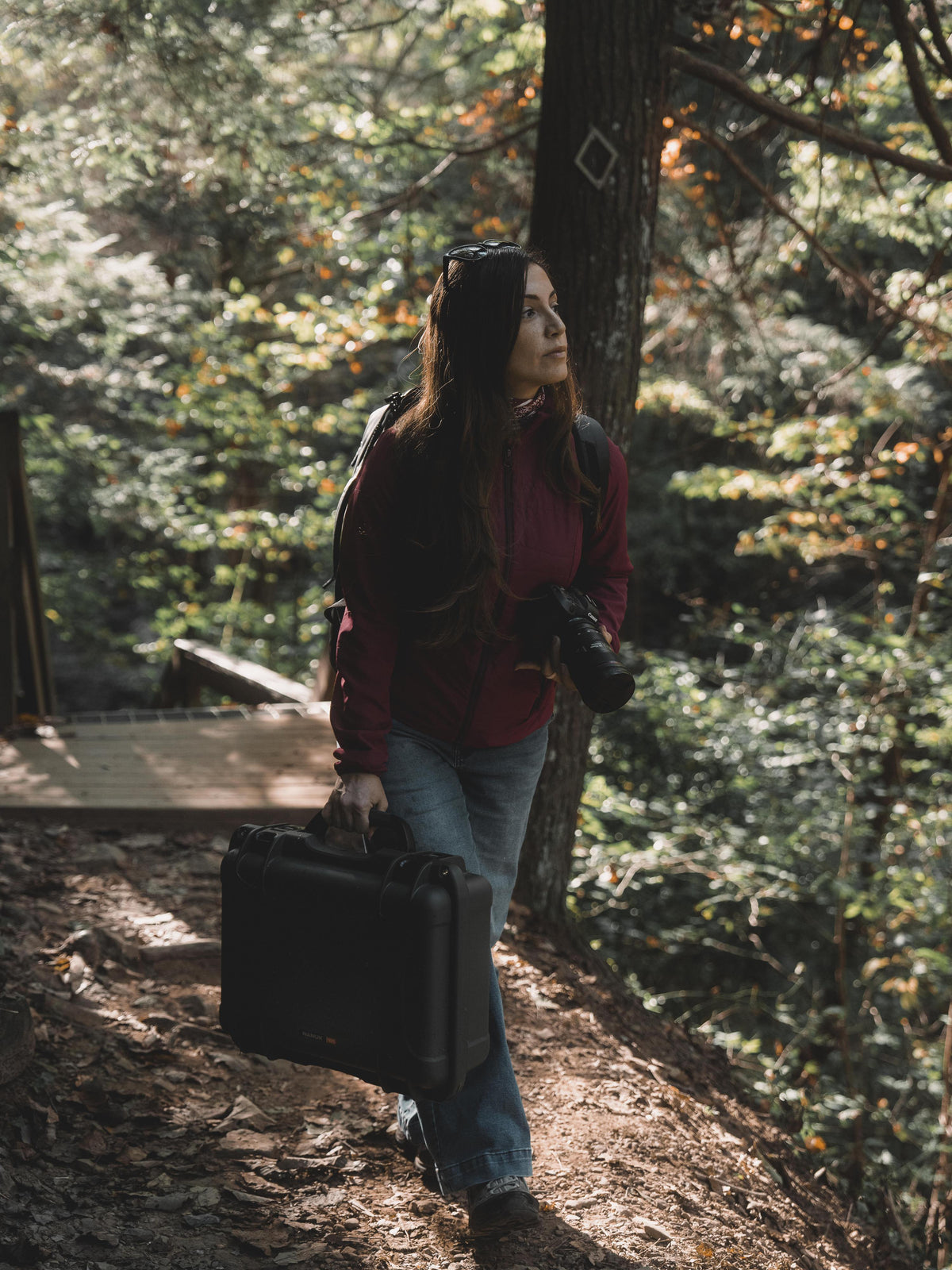 A woman walking on a forest path carrying a black hard-shell briefcase and a camera, wearing a burgundy jacket and blue jeans.