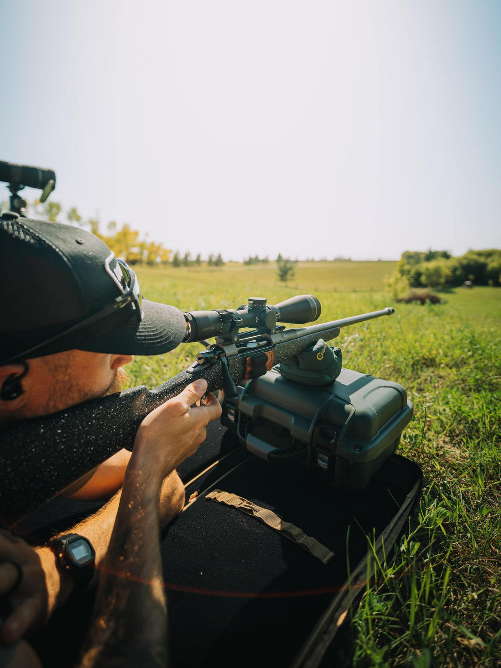 Person aiming a rifle with a scope in a grassy field