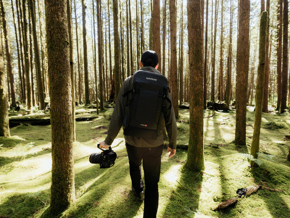 Eine Person, die mit Rucksack und Kamera durch einen Wald geht, umgeben von hohen Bäumen.