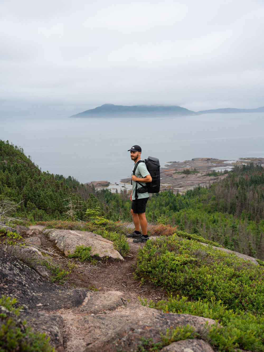 Man with a backpack standing on a rocky outcrop overlooking a scenic landscape.