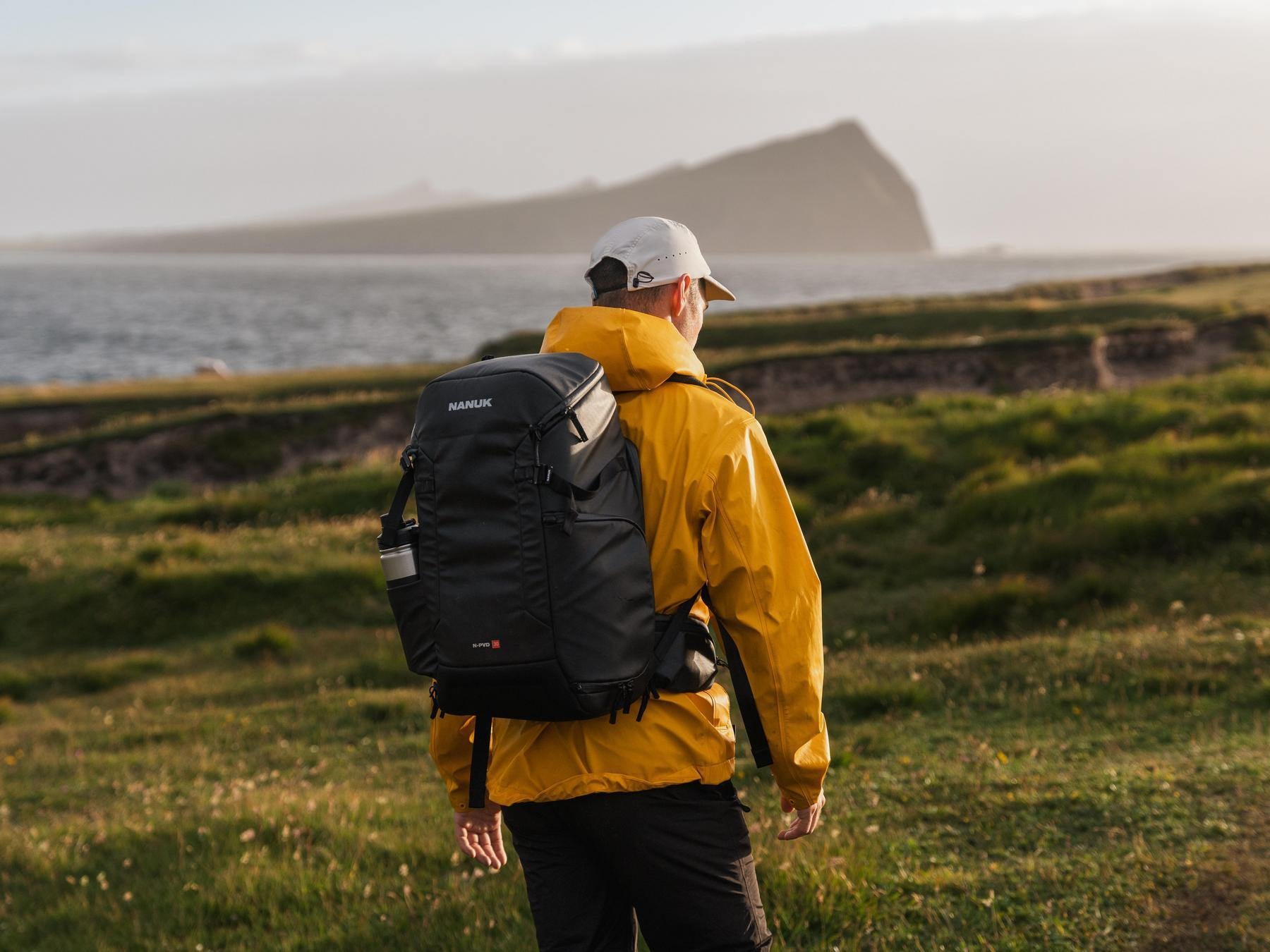 Person wearing a yellow raincoat and black backpack standing on a grassy field with a scenic background.