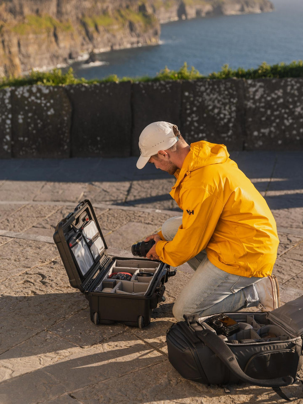 Person in a yellow jacket sitting on a stone ledge with a camera bag and equipment case by the ocean.