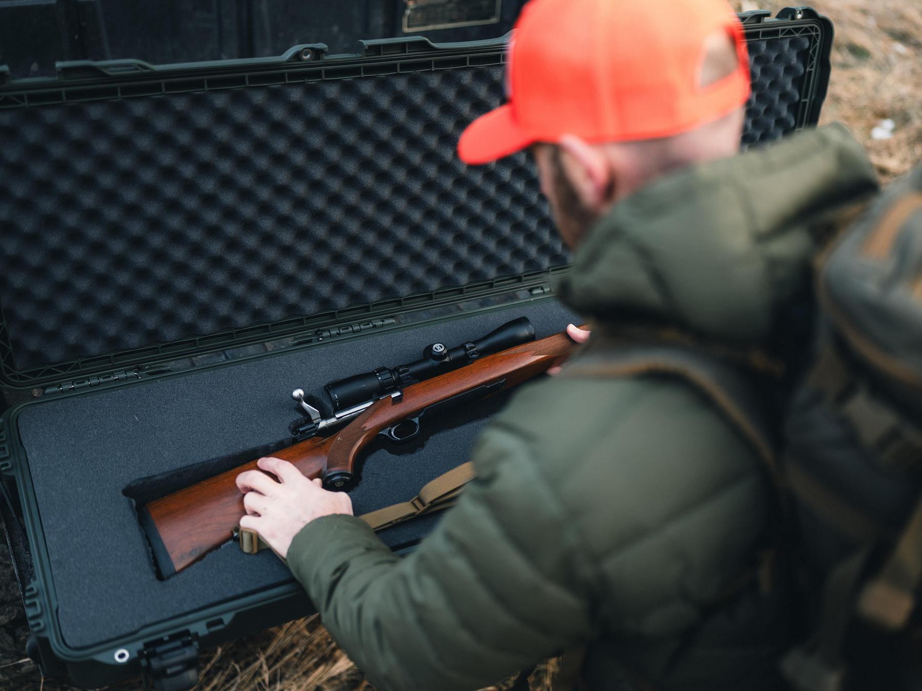 Person handling a rifle in front of an open black rifle case outdoors.