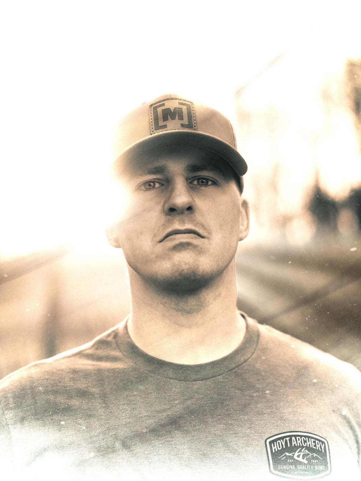Man wearing a cap with a logo and a t-shirt with another logo, standing against a blurred background.