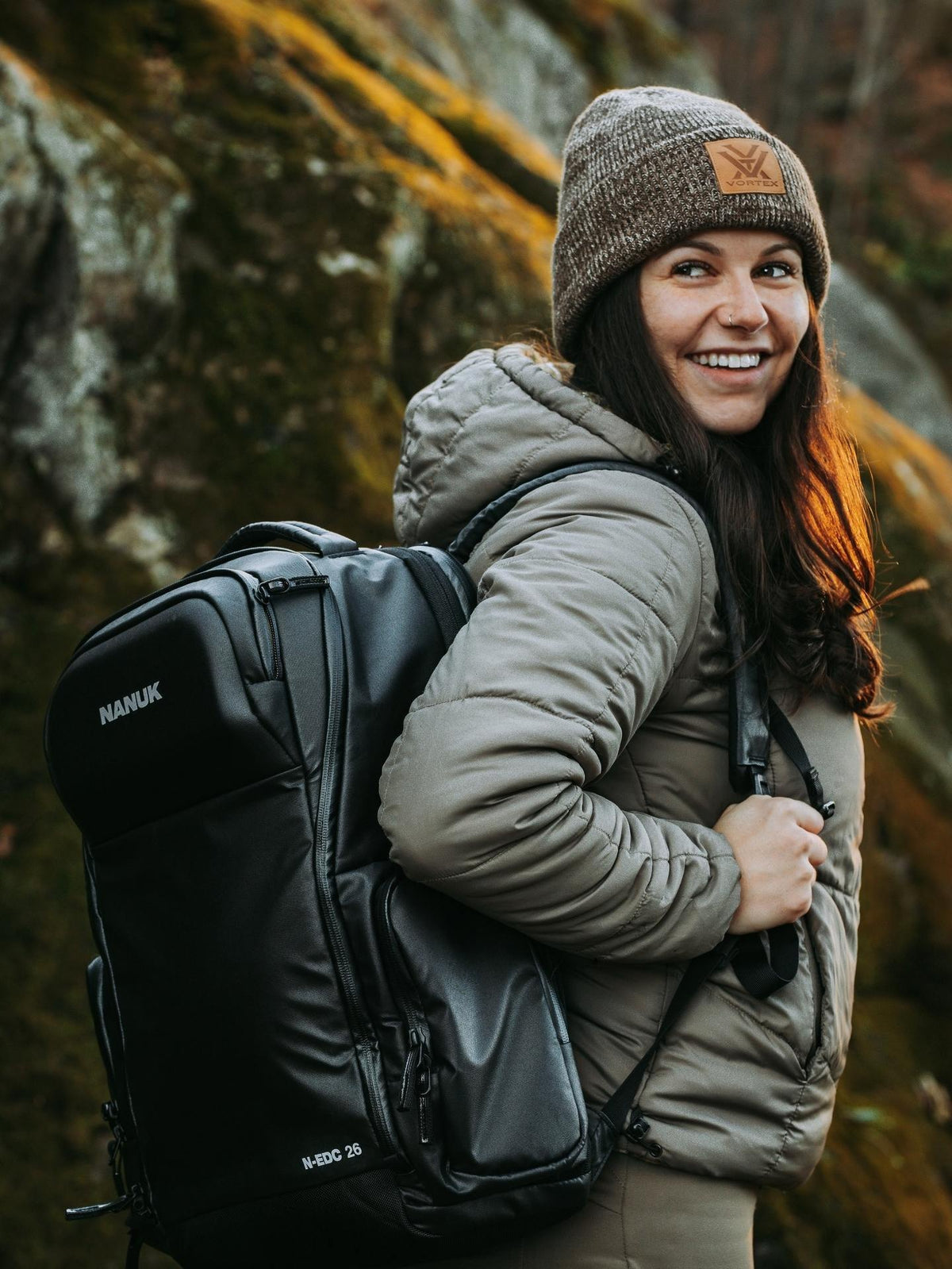 Woman wearing a backpack with a visible brand logo, standing in a natural setting.