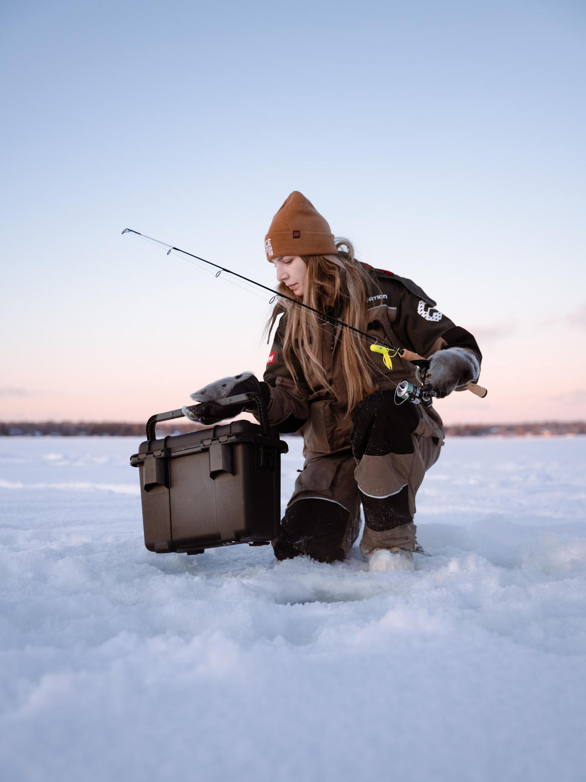 Person ice fishing on a frozen lake with a cooler and fishing gear.