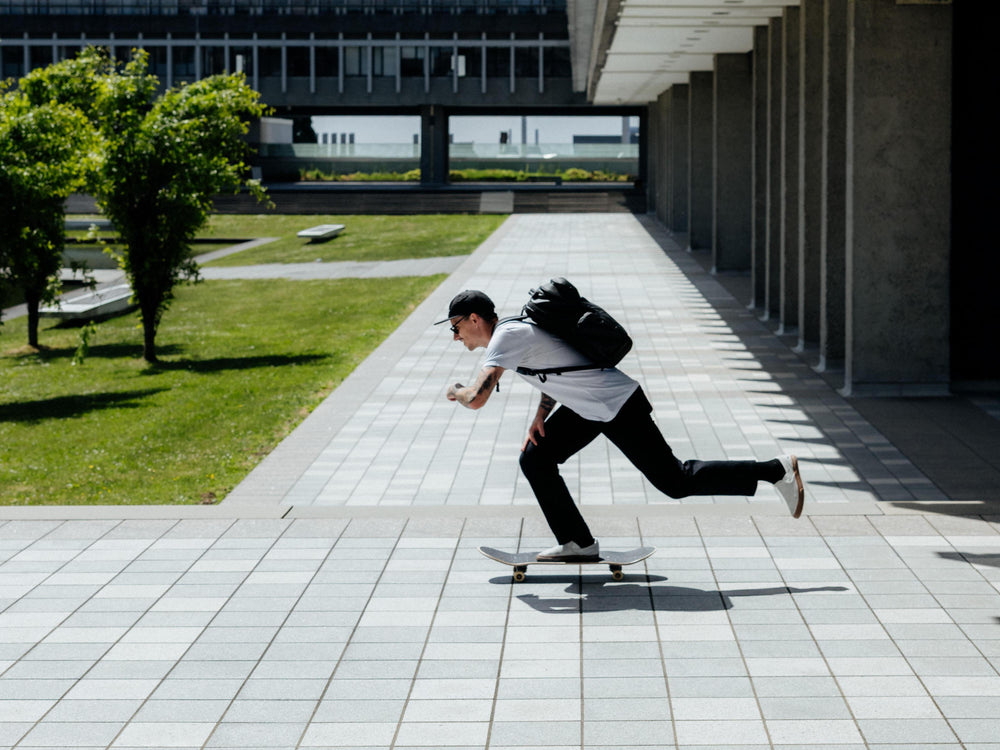 Ein Mann in einem weißen T-Shirt, einer schwarzen Hose und mit einem schwarzen Rucksack fährt auf einem Skateboard über einen modernen Platz im Freien mit Fliesenboden, Rasen und Säulen.
