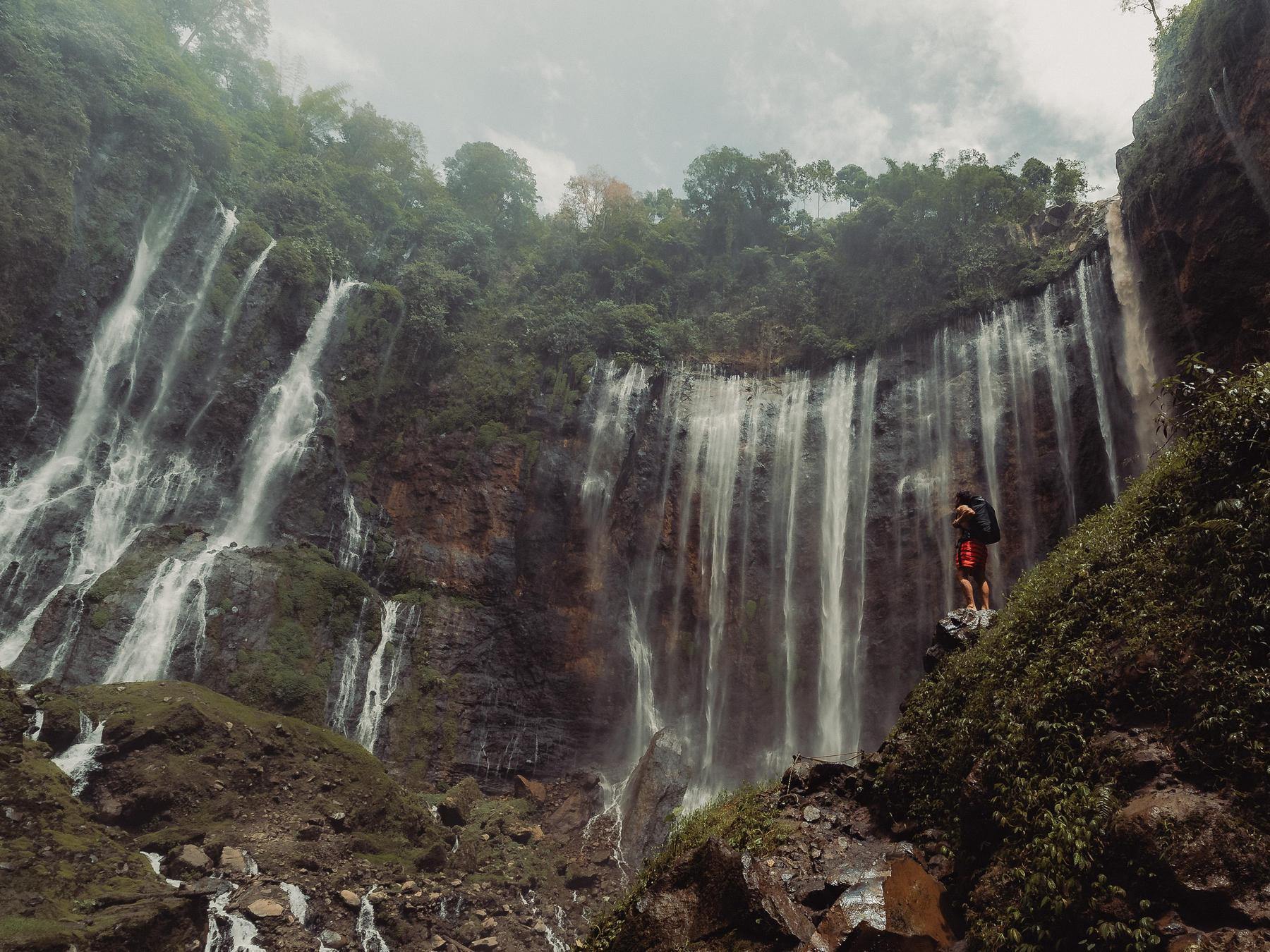 Person standing at the base of a large waterfall in a lush, green forest.