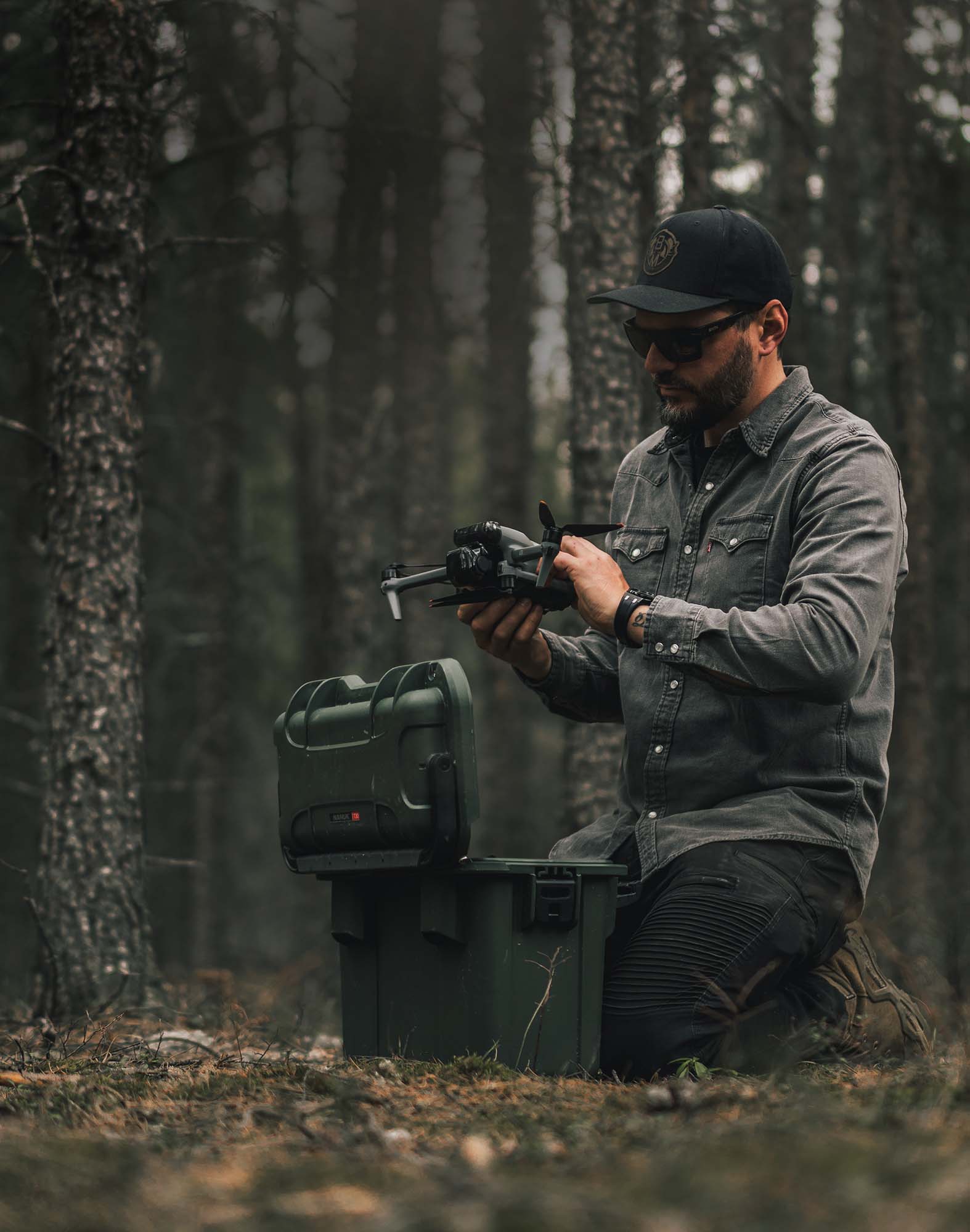 Ein Mann kniet im Wald und bereitet eine Drohne neben einer olivgrünen valise vor.