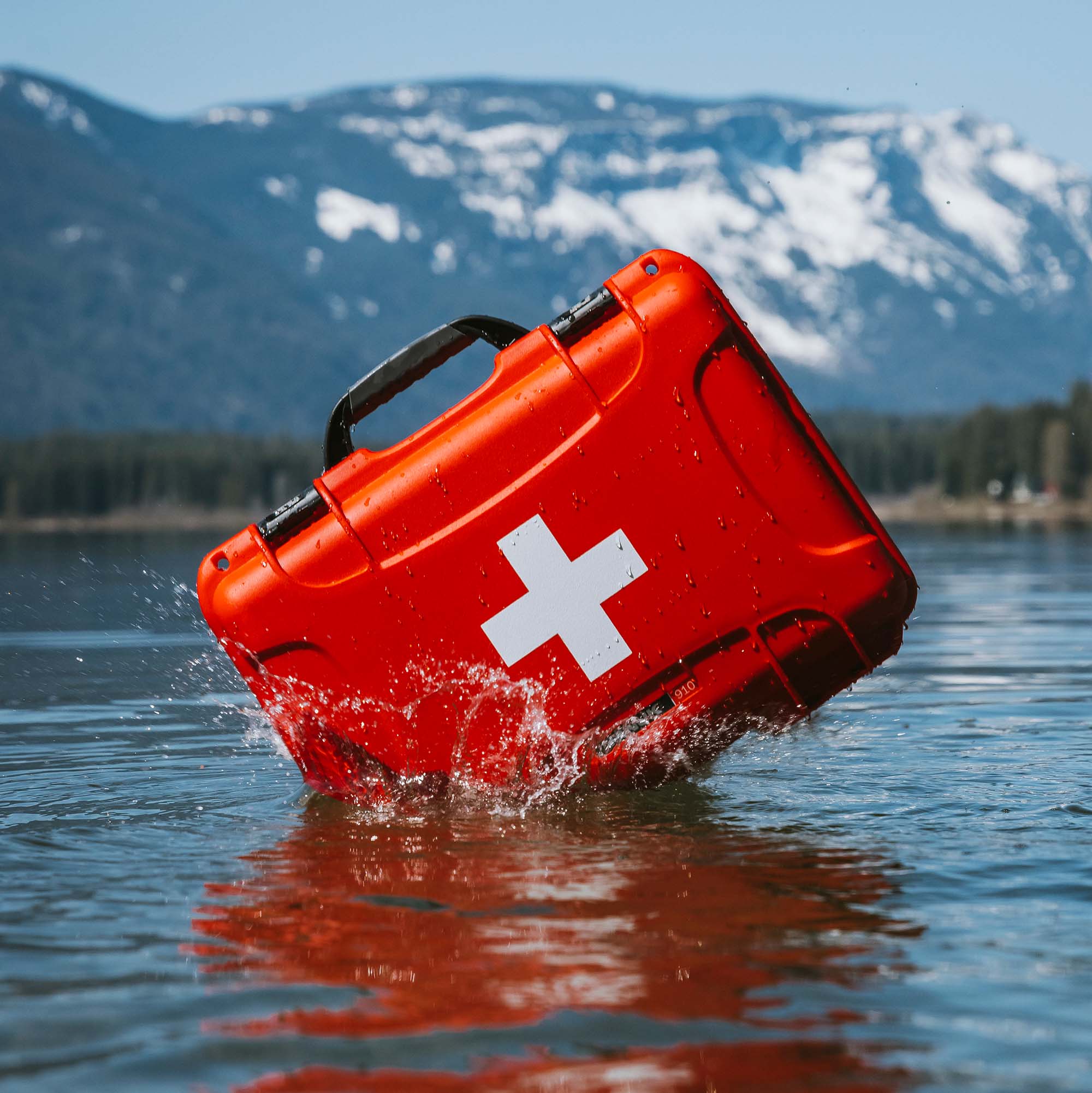 A waterproof red NANUK First Aid Kit floats upright in a calm lake, with snowy mountains in the background.